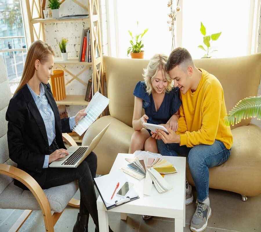 The image shows three people engaged in a collaborative discussion in a well-lit, modern space. One woman, seated in a stylish armchair, is reviewing color swatches and working on a laptop.