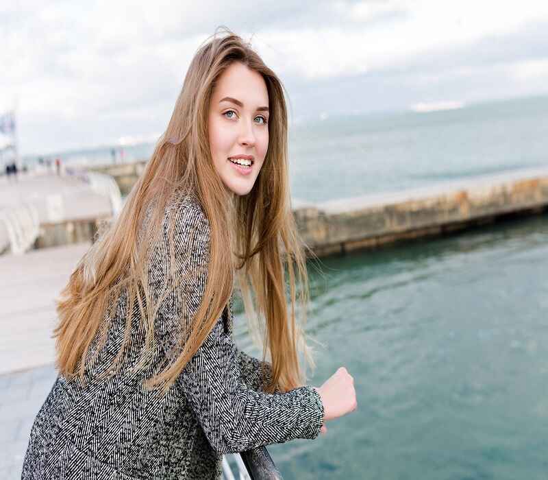 Woman with long brown hair leaning on a railing by the water, wearing a gray blazer and smiling at the camera.