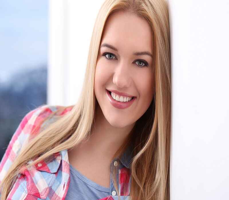 Smiling woman with straight blonde hair wearing a checkered shirt, leaning against a white wall.