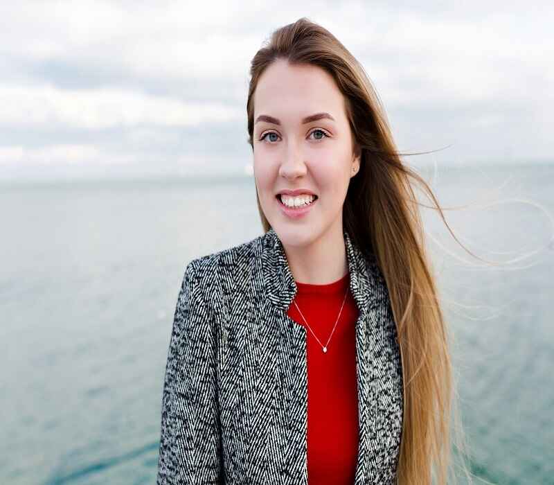 Smiling woman in a gray blazer and red top standing by the ocean with long brown hair flowing in the wind.