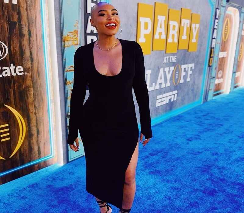 Christine Williamson, ESPN host, dressed in a stylish black slit dress, smiling confidently at an ESPN event backdrop. Known for her bold fashion and unique bald look.