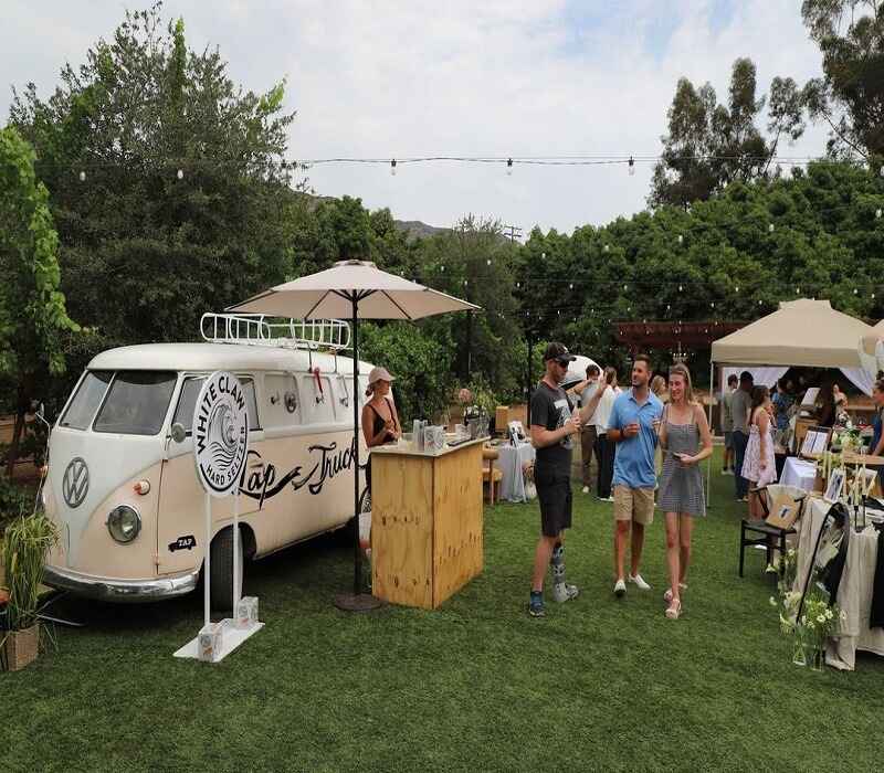 Outdoor event with a vintage White Claw Tap Truck serving drinks, surrounded by people socializing in a garden setting.