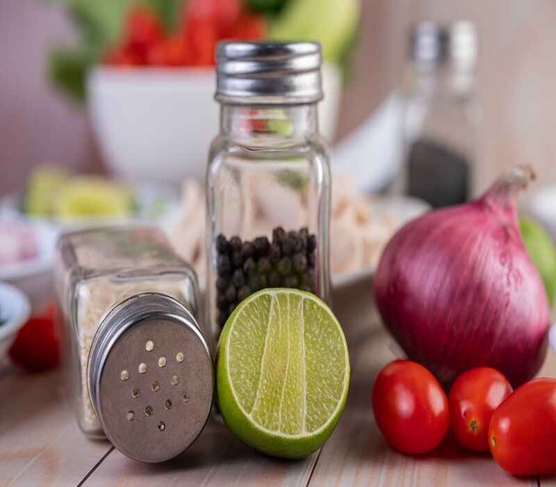 Close-up of kitchen ingredients including a sliced lime, black peppercorns in a shaker, salt shaker, red onion, and cherry tomatoes.