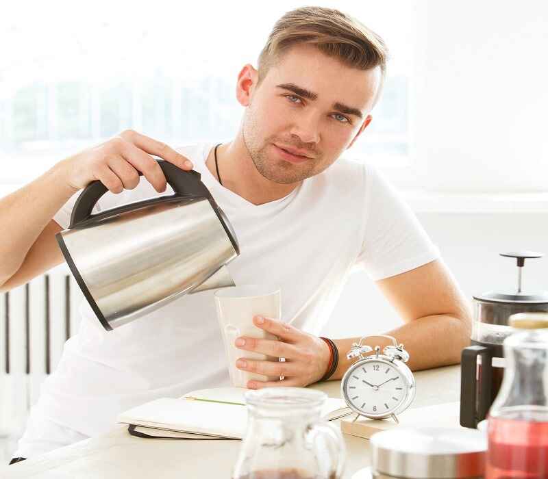 Man in a white shirt pouring hot water into a cup with a kettle, sitting at a table with a notebook, French press, and an alarm clock.