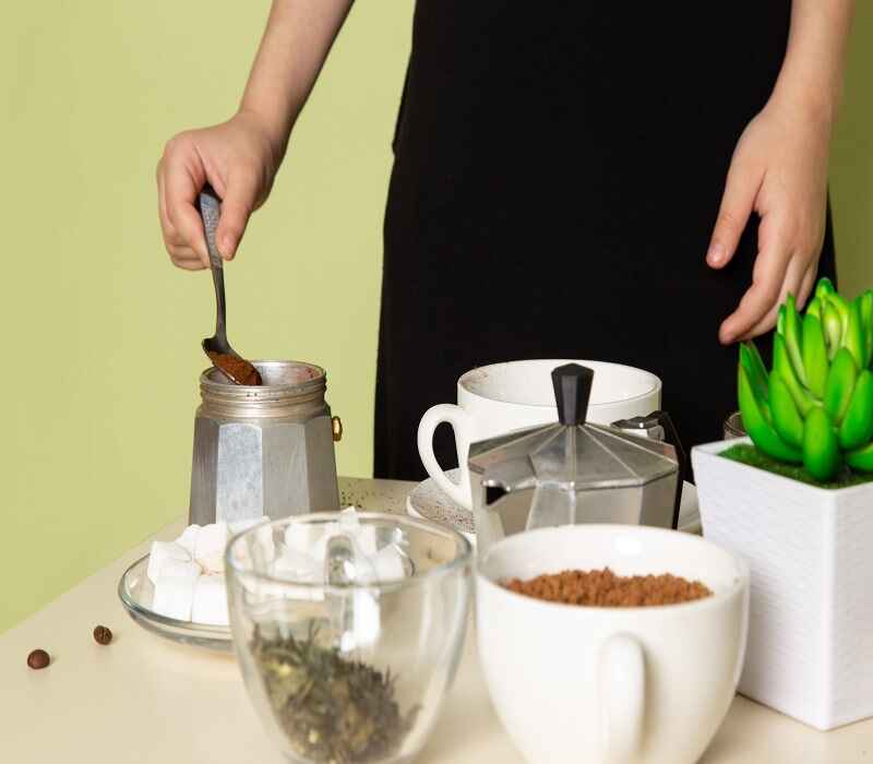 Person preparing coffee using a moka pot, with various ingredients such as ground coffee, sugar cubes, and tea leaves on the table.