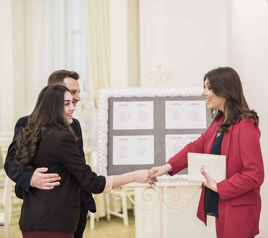 Two professionals warmly greeting each other at a networking event, with a woman in a red blazer shaking hands.