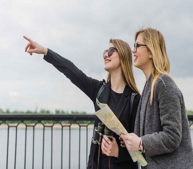 Two young women with a map and binoculars pointing at something in the distance while standing by a river railing.