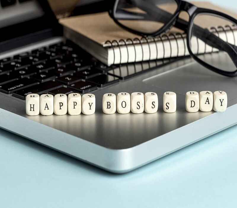 Happy Boss's Day spelled out with letter cubes on a laptop, with a notebook and glasses in the background.