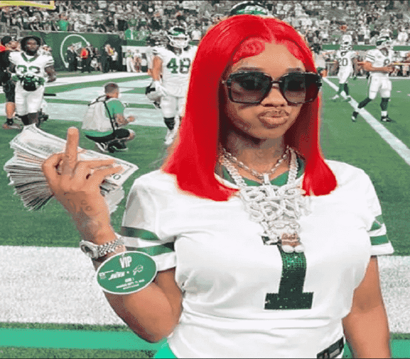 Sexxy Red posing at a football field, holding a stack of cash, wearing a white jersey and bold jewelry, with a confident expression and bright red hair.