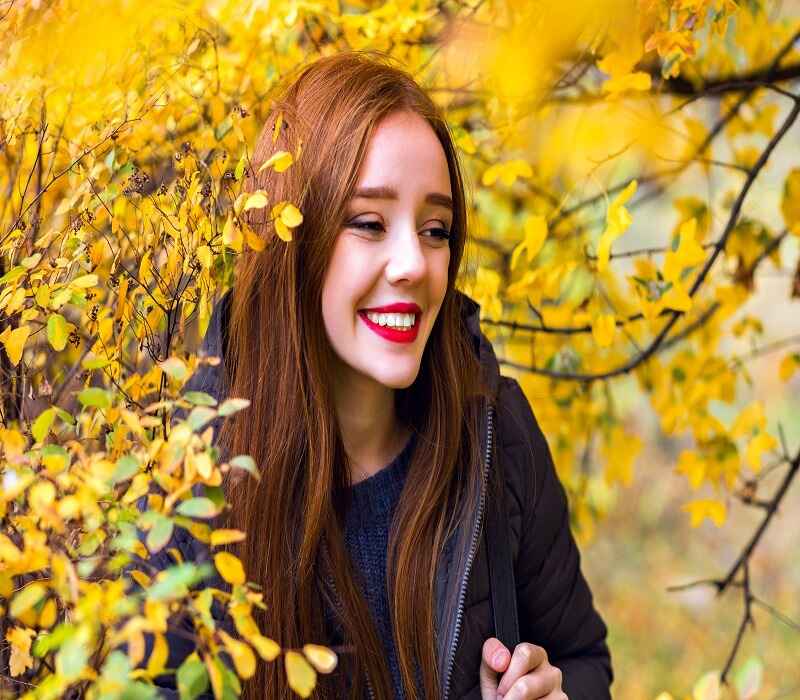 A young woman with long auburn hair and red lipstick smiling brightly amidst yellow autumn leaves.