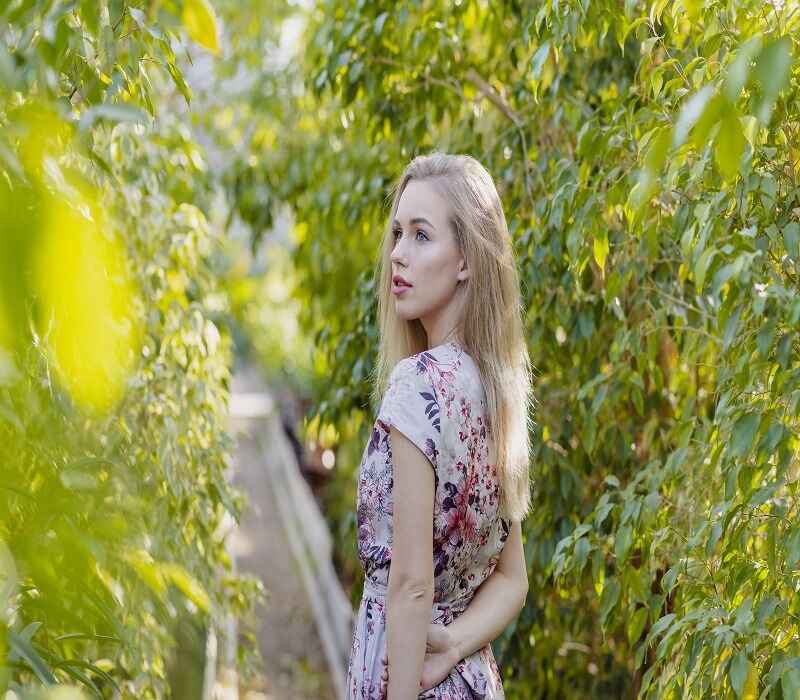 A young woman in a floral dress standing in a green leafy garden, looking over her shoulder with a serene expression.