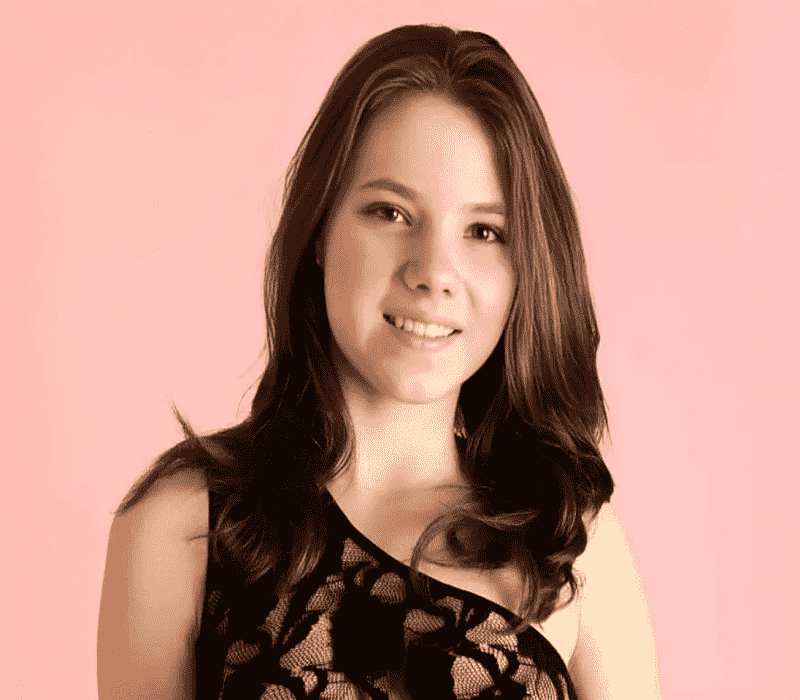 A young woman with shoulder-length dark hair, smiling and wearing a sleeveless black lace dress against a soft pink background.